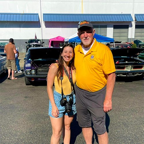 Two people posing together at an outdoor classic car show.