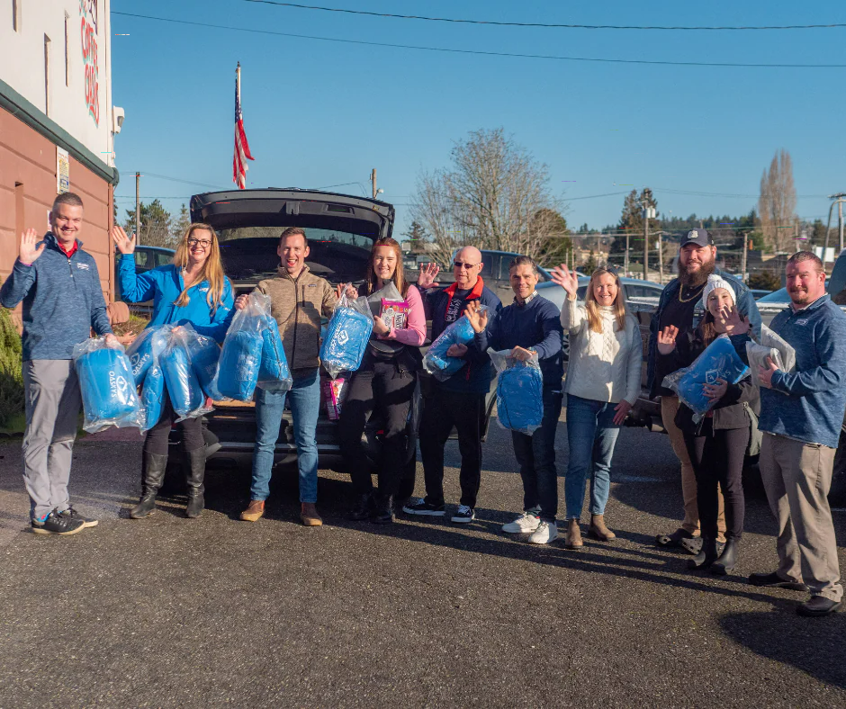A group of people waving and holding blue donation packages behind an open SUV trunk outdoors.