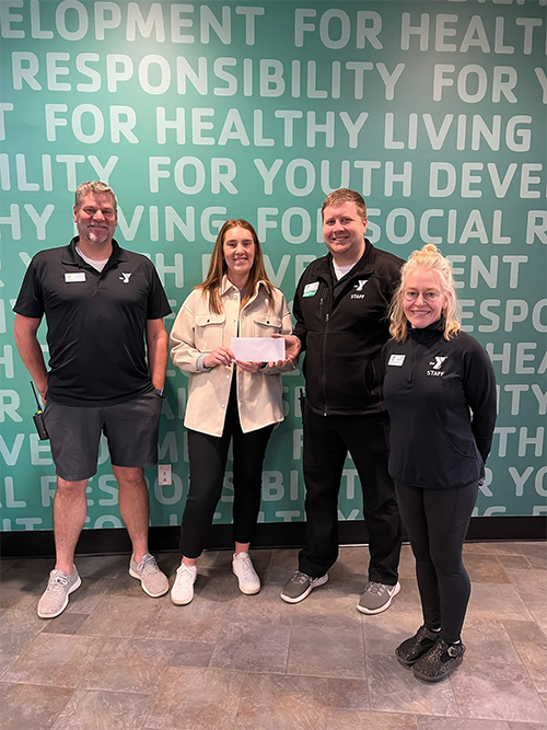 Four individuals posing in front of a teal YMCA wall while the woman in the center holds a white envelope.