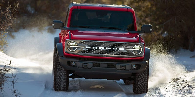 A red Ford Bronco driving through a snowy, wooded landscape, kicking up snow from its tires.