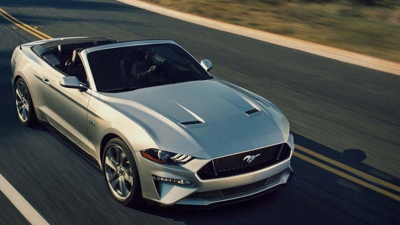 A person drives a silver Ford Mustang convertible on a winding road with the top down.