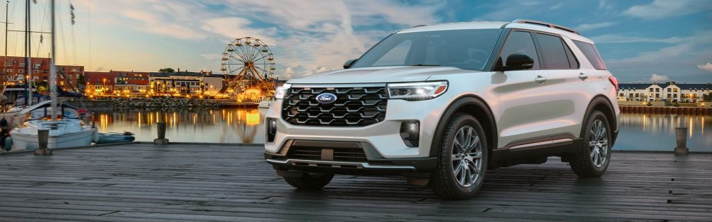 White Ford Explorer SUV on a wooden dock with a waterfront town and ferris wheel in the background at dusk.