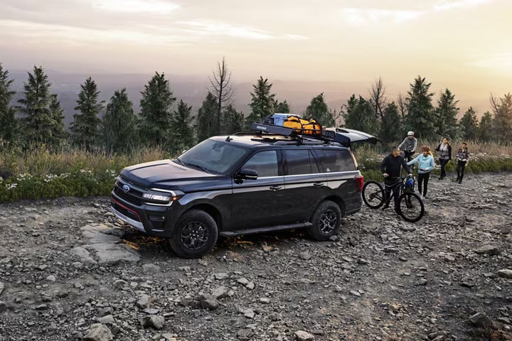 Black SUV with gear on roof rack, parked on a rocky mountain road with a group of people and bikes nearby.