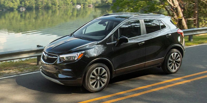 Black Buick Encore SUV driving on a winding, tree-lined road next to a lake with houses in the background.