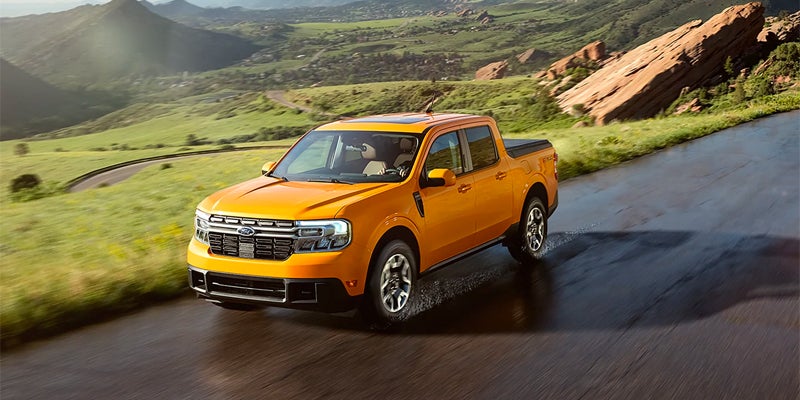 A yellow Ford Maverick pickup truck drives on a winding road through a mountainous, green landscape.