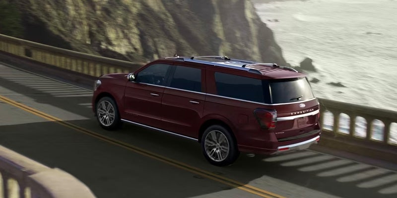 A dark red Ford Expedition SUV driving along a coastal highway with a cliff face on the left and ocean on the right.