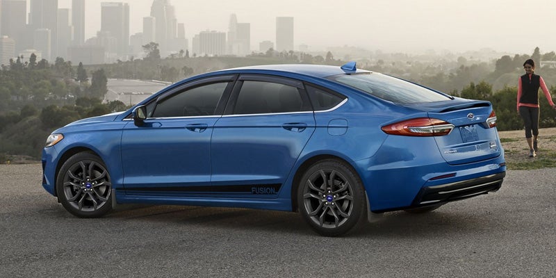 Blue Ford Fusion parked on a hill overlooking a hazy city skyline, with a person walking in the background.