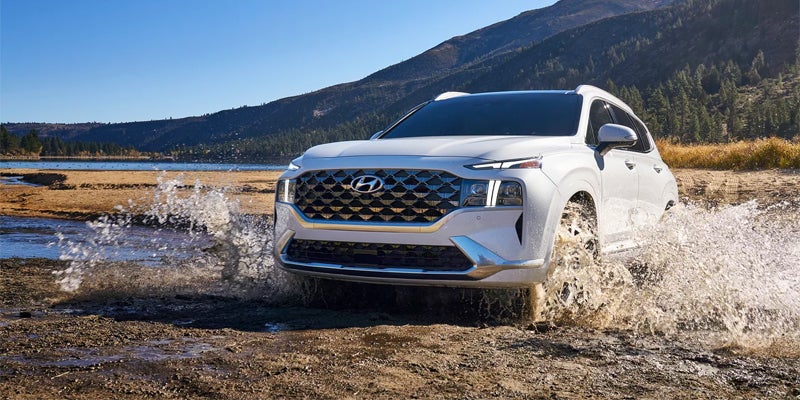 Hyundai Santa Fe driving through shallow water, splashing vigorously, with a lake and mountains in the background.