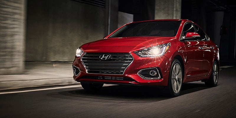 Hyundai Accent sedan with glowing headlights drives left on a dark road in front of blurred concrete.