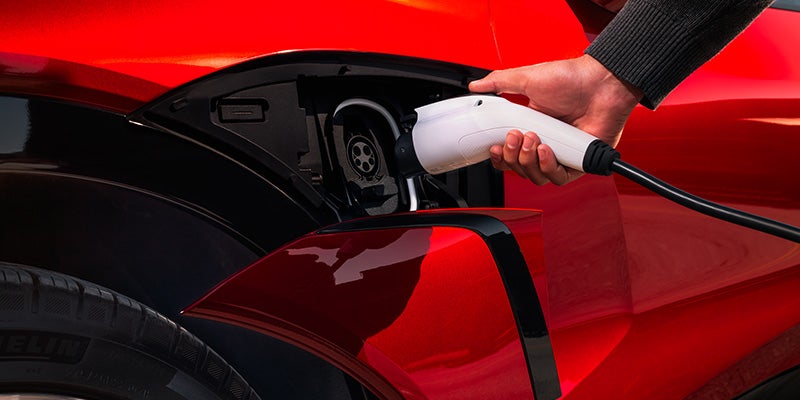 A person's hand inserts a white electric vehicle charging cable into the charge port of a red car