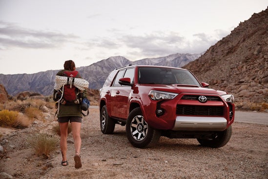 Red Toyota 4Runner SUV with rooftop cargo on a dirt road, mountains in the background.