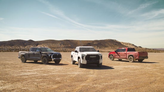 Three Toyota Tacoma pickup trucks, one black, one white and one red, parked on a dirt road with mountains and stars at dusk.