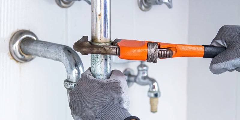 Gloved hands using an orange pipe wrench to tighten a chrome pipe, part of a sink plumbing system.