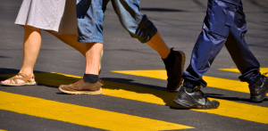 Three people walking across a yellow-striped crosswalk on a sunny day, highlighting pedestrian safety awareness