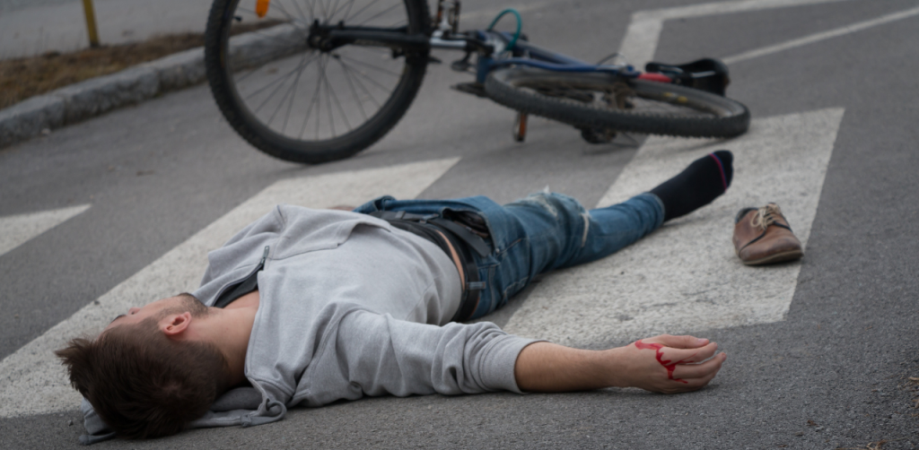 Injured cyclist lies on a crosswalk near a fallen bike, with blood on his hand, showing pedestrian accident aftermath