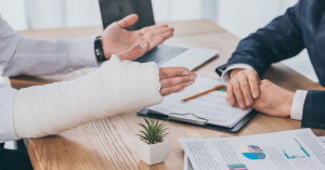 A person with a bandaged hand discusses paperwork with another person in a suit, likely a lawyer, at a desk with documents and a small plant