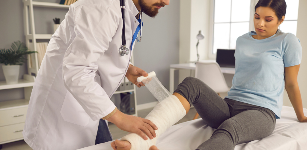 A doctor wrapping a bandage around a woman's leg in a medical setting, with the woman sitting on an exam table and looking down at her leg