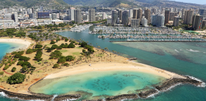 Aerial view of coastal city with turquoise lagoon, sandy beach, marina, and high-rise skyline against green mountains