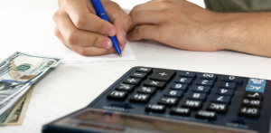 Person writes on paper next to a stack of hundred-dollar bills and a calculator, symbolizing financial planning or evaluating a personal injury settlement amount