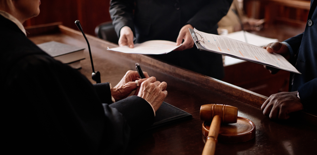Judge signs legal documents at courtroom bench, with lawyers presenting paperwork and gavel visible