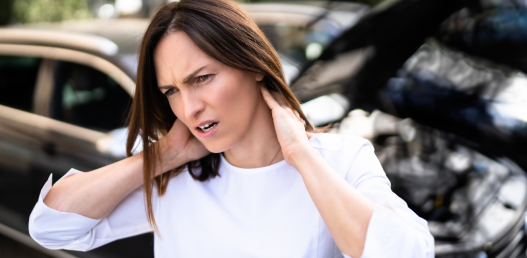 A woman touching her neck with both hands, appearing to be in pain, standing in front of a car with its hood open, possibly after an accident