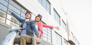 Smiling couple riding a scooter with arms outstretched, and a parked yellow scooter on a city street near a concrete wall