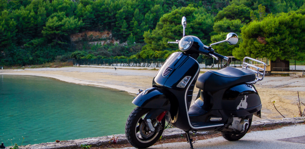 Black scooter parked by a scenic beach with clear water and lush greenery in the background