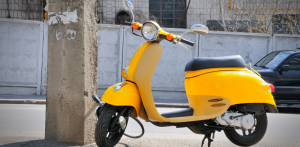 Yellow scooter parked on a city street beside a concrete post, with urban buildings and a car in the background