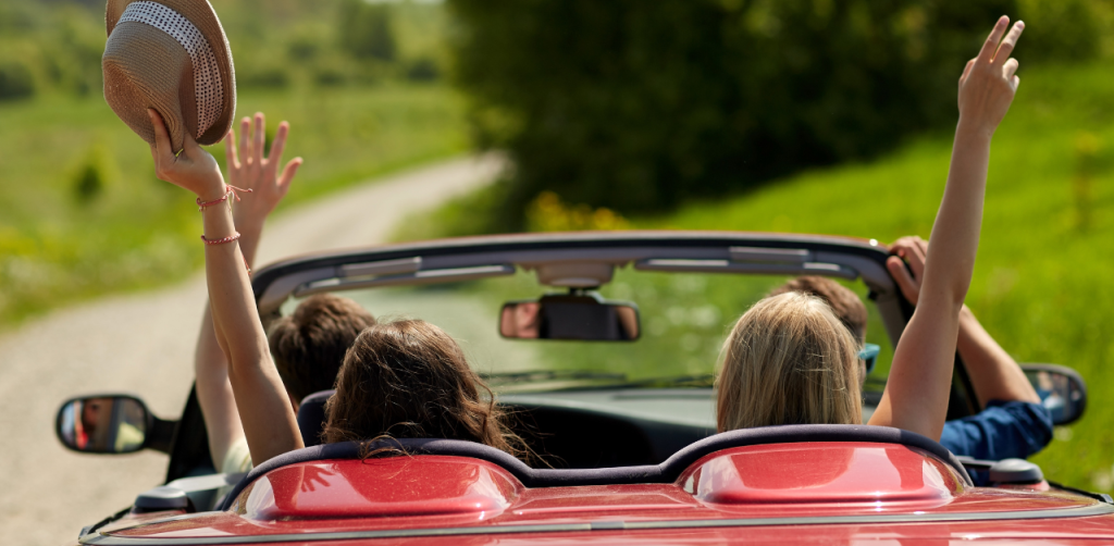 Group of friends in a red convertible, arms raised, enjoying a carefree road trip on a scenic countryside road