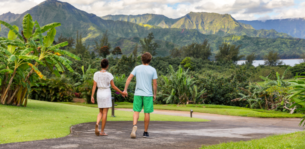 A couple walking hand in hand on a lush green pathway surrounded by tropical plants, with a backdrop of towering green mountains under a partly cloudy sky