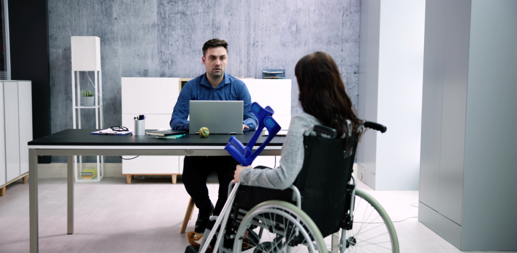 Man in a blue shirt sits at a desk across from a woman in a wheelchair, holding crutches, in a modern office setting, engaging in conversation