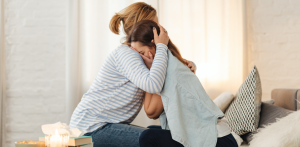 Two women embrace in a comforting hug, one visibly emotional on a living room couch
