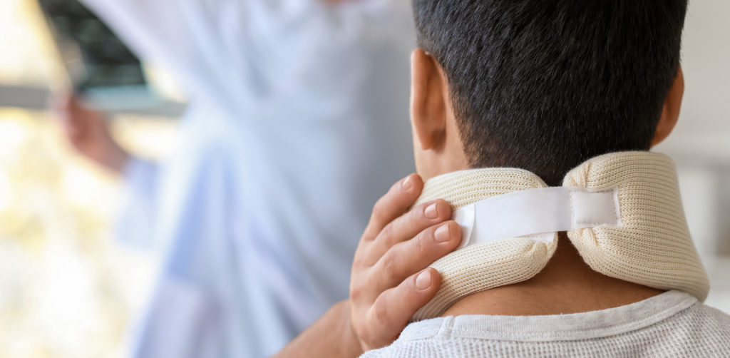 A man wearing a neck brace is seen from behind, holding his neck while a doctor reviews an X-ray nearby