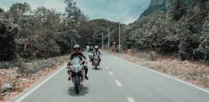 Group of motorcyclists wearing helmets ride along a forested road with power lines and traffic signs