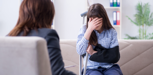 Injured woman with arm in sling and crutch, covering her face while speaking with a professional