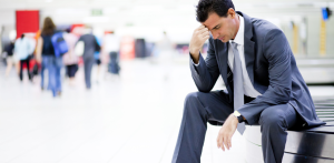 A man in a suit sitting at an airport, holding his head in frustration, with blurred travelers in the background