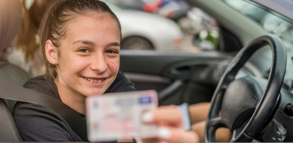Smiling teen in a car proudly holds up a new driver's license, excited about earning the privilege to drive