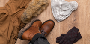 Winter clothing essentials including a brown parka with fur hood, leather boots, gray knit beanie, and dark gloves arranged on a wooden floor