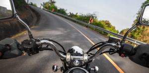 First-person view riding a motorcycle on a winding road, handlebars visible with guardrails and greenery ahead