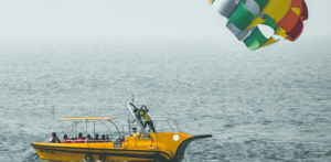 A bright yellow boat tows a colorful parasail over open water, with people onboard watching