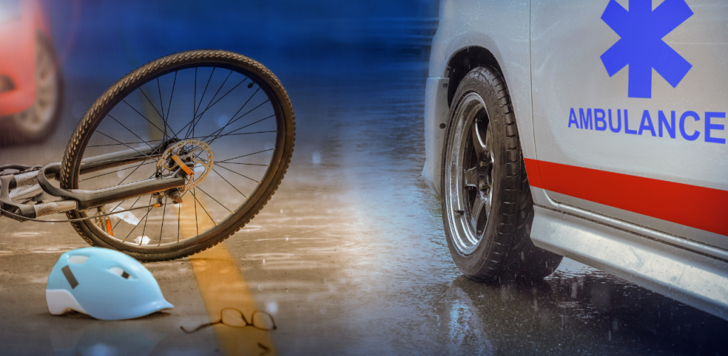 Fallen bicycle, blue helmet, and glasses on a wet road beside an ambulance, indicating a recent accident in rainy weather