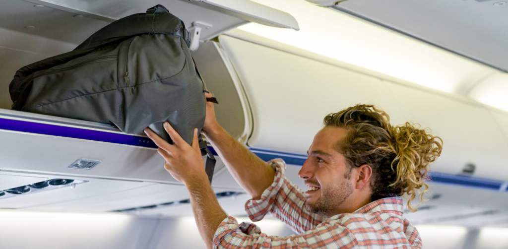 Smiling man places a backpack into the overhead bin on an airplane, following airline safety regulations for carry-on luggage