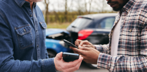 Two drivers using smartphones to exchange information after a car accident, with damaged vehicle visible in the background