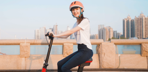 Smiling woman in helmet rides an electric scooter along a waterfront path with a city skyline in the background