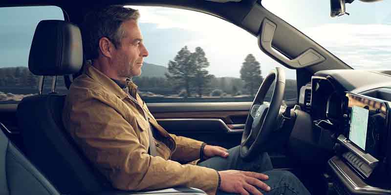 Middle-aged man in a brown jacket looks out the window from the driver's seat of a car, a landscape passes by.
