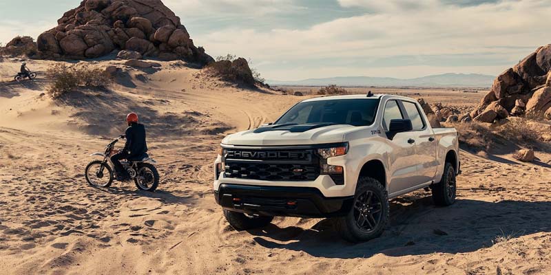 White Chevrolet pickup truck in a sandy desert with rock formations, a person on a dirt bike in the background.