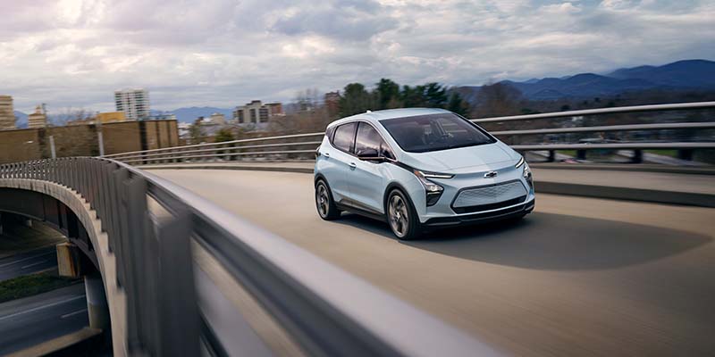 Light blue Chevy Bolt EV driving on a curved city overpass with buildings and mountains in the background under a cloudy sky.