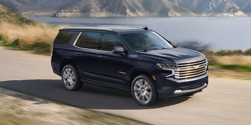 Dark blue Chevrolet Tahoe SUV driving on a winding road next to a lake with mountains in the background.