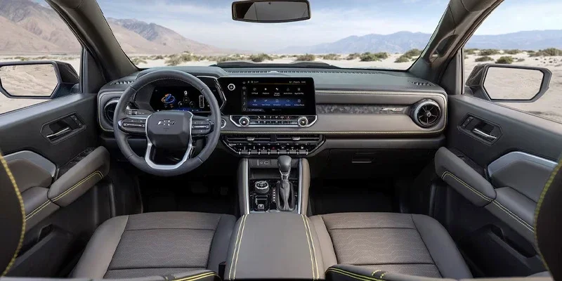 Interior of a car with a black steering wheel, digital displays, and black seats with yellow stitching, looking out at a desert.