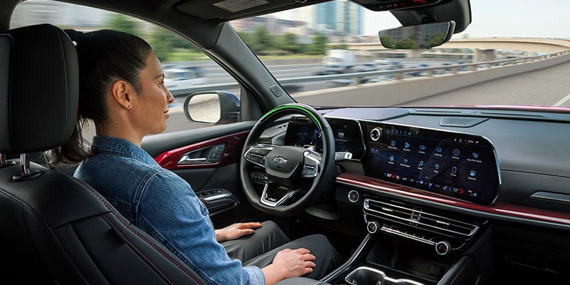 Woman in driver's seat of modern car on highway, looking ahead. Dashboard features large screens and a glowing green steering wheel.